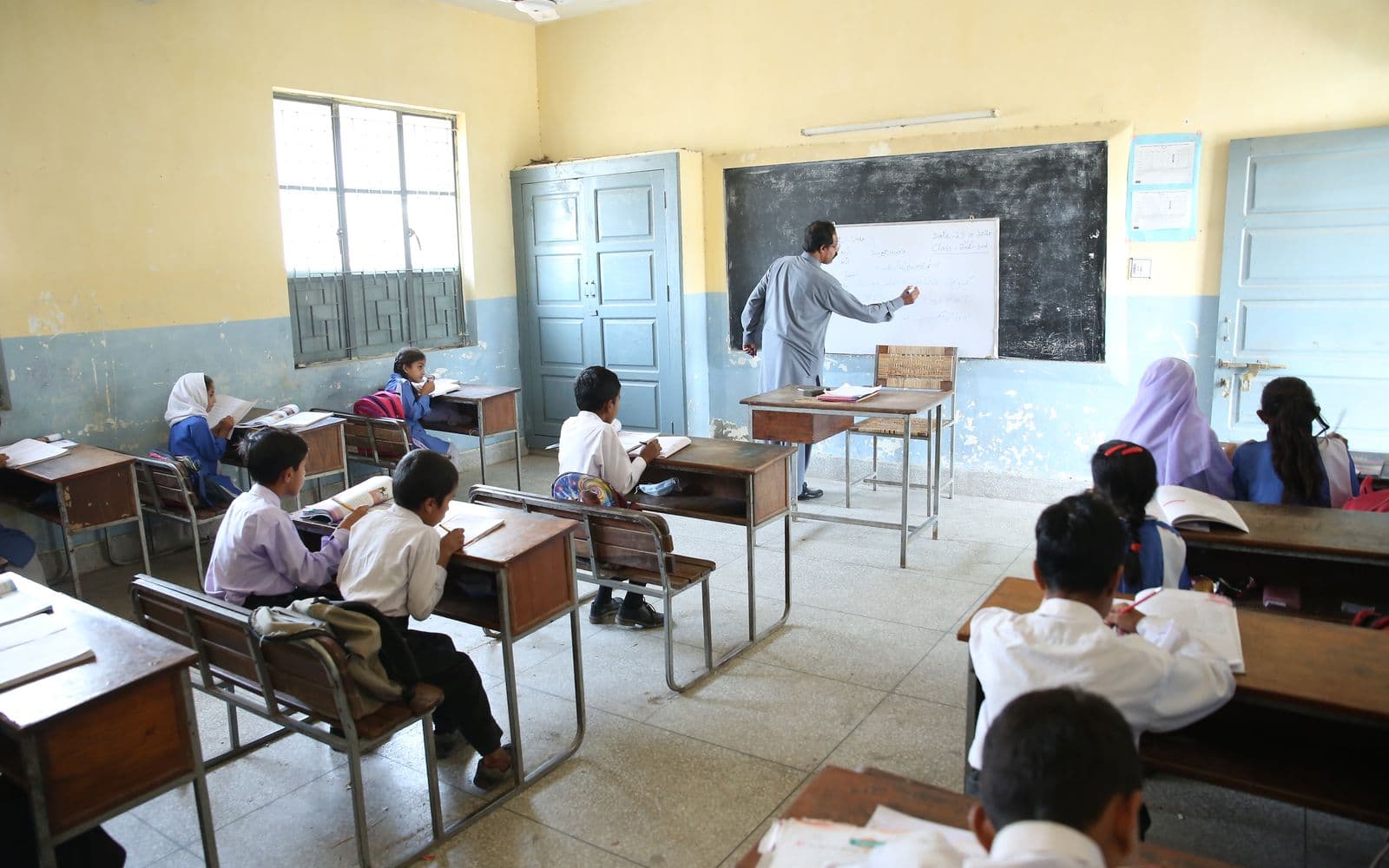 Pakistani classroom with students at wooden desks and a teacher writing on the blackboard